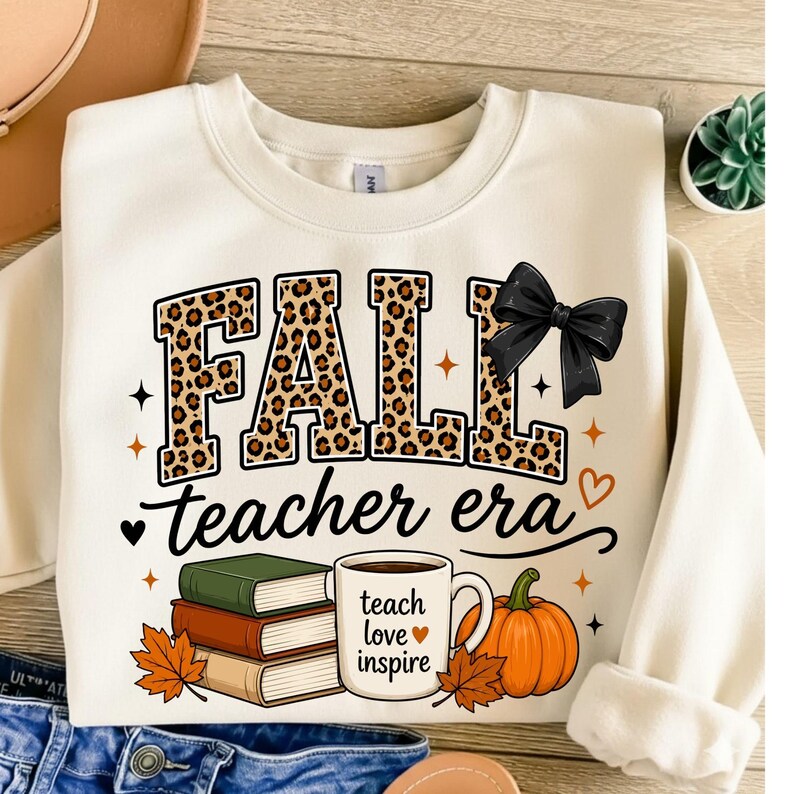 Puede incluir: Una sudadera de cuello redondo color crema con el texto FALL en estampado de leopardo con un lazo negro, y el texto teacher era debajo. El dise&ntilde;o incluye una pila de tres libros, una taza con el texto teach love inspire y una calabaza.