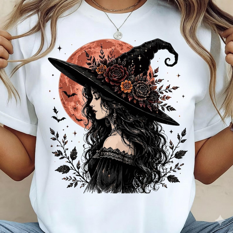 May include: A white t-shirt featuring a graphic illustration of a person with long dark hair wearing a black witch hat adorned with dark red and orange roses. The background shows a large orange moon, flying bats, and dark leafy branches.