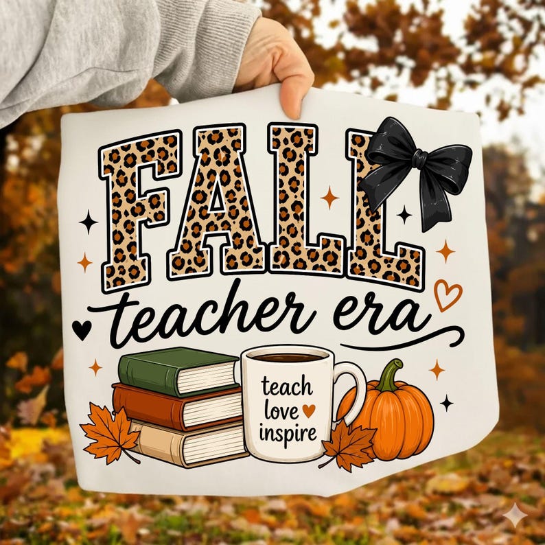 Puede incluir: Una camiseta blanca con el texto FALL en estampado de leopardo con un lazo negro, y el texto teacher era debajo. Incluye una pila de tres libros, una taza blanca con el texto teach love inspire y una calabaza de color naranja.