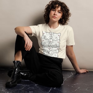May include: Cream-colored t-shirt featuring a stack of cartoon cat faces with black outlines and pink accents. The shirt is made of a soft material. The model is wearing black pants and black boots.