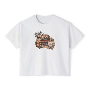 Peut inclure: Un t-shirt blanc à manches courtes orné d'un motif représentant un chat tigré endormi sur une pile de livres, une bougie allumée, des lunettes, une tasse avec le texte Books Coffee Happiness et une écharpe rayée, entouré de petites fleurs.