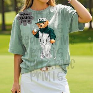 May include: A light green t-shirt featuring a cartoon bear dressed in a golf outfit, holding a golf ball and club. The bear wears a green and white jacket, white pants, and a cap with "AUG" on it. The shirt also has the text "instant download png file".
