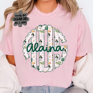 May include: A pale pink t-shirt with a circular design featuring the name "Alaina" in green script. The design incorporates a monogram with a gingham pattern and illustrations. The text on the shirt says "ready for DOWNLOAD in 1-3 days!"