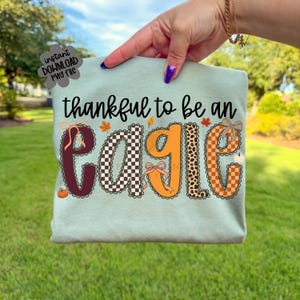 May include: A light green t-shirt featuring the black text "thankful to be an eagle" with the word "eagle" in large, patterned letters. The letters include maroon, black and white checkerboard, orange, leopard print, and orange checkerboard patterns.