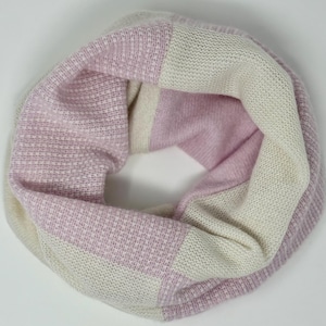 May include: A knitted infinity scarf in a soft color palette of pale pink and off-white. The scarf features a striped pattern with alternating blocks of color. The fabric appears to be a soft, textured knit, suitable for cooler weather.
