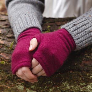 May include: A pair of magenta fingerless gloves are worn under a gray knit sweater. The gloves are made of a soft material and cover the hand up to the knuckles, leaving the fingers free. The sweater has ribbed cuffs.