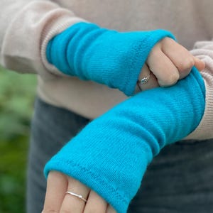 May include: A pair of bright turquoise arm warmers. The warmers are made of a soft, ribbed knit material. The person is wearing a light pink sweater and gray pants. The arm warmers are a vibrant pop of color.