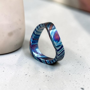 Könnte beinhalten: Ein einzigartiger Ring mit einem farbenfrohen, marmorierten Design. Der Ring hat Schattierungen von Blau, Lila und Weiß und eine markante, eckige Form. Der Ring ist ein Schmuckstück, wahrscheinlich für den täglichen Gebrauch.