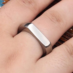 May include: A silver-colored ring with a rectangular design. The ring is worn on a finger and has a smooth, polished surface. The ring is a simple, modern piece of jewelry.