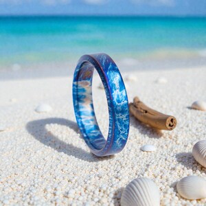May include: A blue ring with a wave-like pattern, resting on white sand with seashells and a piece of driftwood. The ring's design resembles ocean waves, with shades of blue and white. The background features a turquoise ocean and blue sky.