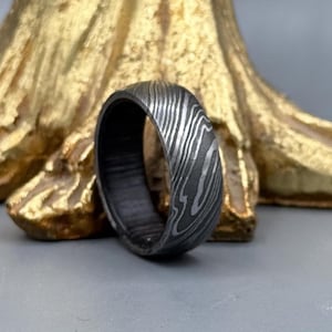 Pode incluir: Um close-up de um anel de aço Damasco. O anel tem um interior escuro e texturizado e um padrão de redemoinho de metal claro e escuro no exterior. O anel é colocado contra um fundo dourado desfocado.