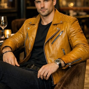 Puede incluir: Chaqueta de cuero amarillo mostaza con camiseta negra y vaqueros oscuros. La chaqueta presenta un estilo motero clásico con cuello de solapa, cremallera asimétrica y bolsillos con cremallera. Se ve un reloj de pulsera y una pulsera.