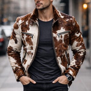 May include: A brown and white cowhide leather jacket with a zip-up front and a fur collar. The jacket has two zippered pockets and is worn with a black t-shirt and black jeans. A watch is visible on the wrist.