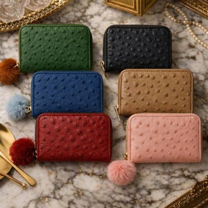 Könnte beinhalten: Sechs kleine, rechteckige Geldbörsen in verschiedenen Farben: Grün, Schwarz, Blau, Beige, Rot und Rosa. Jede Geldbörse hat eine strukturierte Oberfläche, einen goldenen Reißverschluss und einen flauschigen Pompon. Auf einer Marmoroberfläche angeordnet.