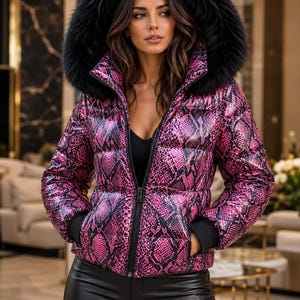 May include: A puffer jacket featuring a vibrant pink and black snakeskin pattern, a high collar, and a large black faux fur-trimmed hood. The jacket has a front zipper closure, long sleeves with black ribbed cuffs, and a quilted, insulated design.