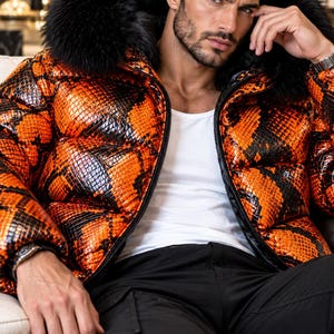 Könnte beinhalten: Eine Steppjacke mit einem leuchtend orangefarbenen und schwarzen Schlangenmuster, einem Frontreißverschluss und einem großen schwarzen Kunstfellbesatz an der Kapuze. Die Jacke wird über einem schlichten weißen Unterhemd und einer schwarzen Cargohose getragen.
