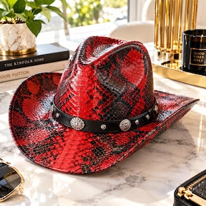 Puede incluir: Un sombrero de vaquero con estampado de piel de serpiente en rojo y negro, que presenta una banda negra adornada con tachuelas circulares plateadas y conchos metálicos en forma de flor. El sombrero tiene una copa estructurada y ala ancha curva.