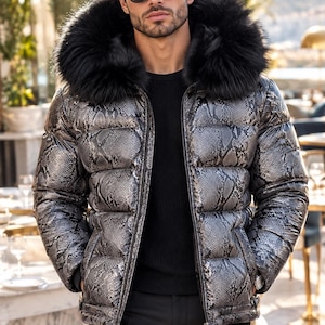 Pode incluir: Um casaco acolchoado com padrão de pele de cobra prateado e preto, fecho de correr frontal preto e um capuz grande com rebordo de pelo sintético preto. O casaco é usado sobre uma camisola preta com calças pretas e óculos de sol escuros.