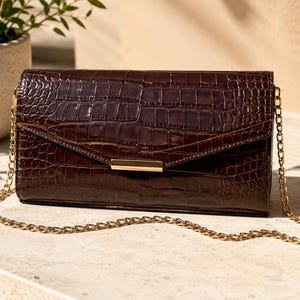 May include: A dark brown rectangular clutch bag with a glossy crocodile skin texture. The bag features a pointed envelope-style flap closure with a horizontal gold-tone metal accent bar and a thin gold-tone metal chain shoulder strap.