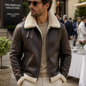 May include: A dark brown leather bomber jacket featuring a thick cream-colored shearling collar and matching shearling trim at the cuffs and hem. The jacket includes a front zipper closure, two side pockets, and two small brown leather buckle straps.