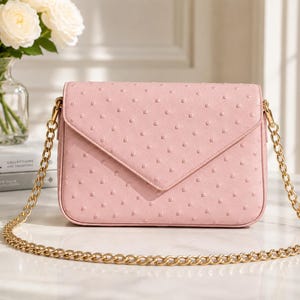 Puede incluir: Un bolso de mano rosa claro rectangular con cierre de solapa y cadena dorada. El bolso tiene una superficie texturizada con pequeños puntos espaciados. La cadena es de color dorado brillante. El bolso está sobre una superficie blanca.