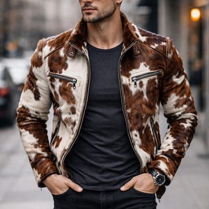 May include: A brown and white cowhide leather jacket with a zip-up front and a fur collar. The jacket has two zippered pockets and a brown leather trim. The person is wearing a black t-shirt and black jeans.