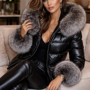 Könnte beinhalten: Schwarze Steppjacke mit glänzendem Finish und Pelzbesatz an Kapuze und Manschetten. Dazu schwarze Lederhose. Die Person trägt Goldschmuck und eine Armbanduhr.