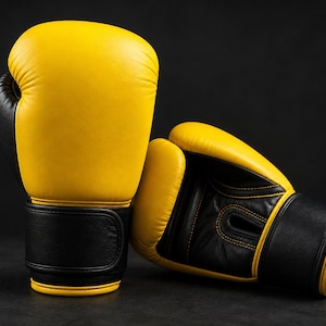 May include: A pair of yellow and black boxing gloves. The gloves are made of leather and have a classic design. The gloves are on a dark background, and the lighting highlights the texture and shape of the gloves.