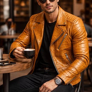 Puede incluir: Chaqueta de cuero marrón claro con textura, cierre de cremallera y cuello clásico. Combinada con una camiseta negra y vaqueros oscuros. La persona lleva gafas de sol y un reloj. La imagen está tomada en un café. La chaqueta es color caramelo.