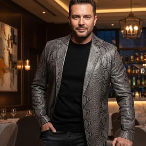 May include: A grey snakeskin-effect blazer worn over a black top and dark jeans. The blazer has a textured finish and is worn in a restaurant. The person is standing with one hand in their pocket.