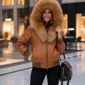 Peut inclure: Veste en cuir marron avec une capuche bordée de fourrure. La veste de style bombardier a une fermeture éclair et des poignets et ourlets côtelés. Un sac à main noir est porté. Le cuir semble texturé.