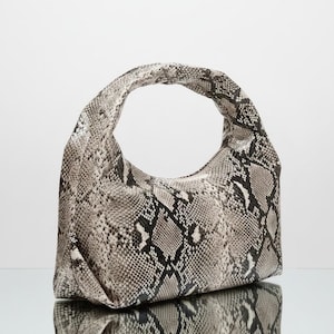 Peut inclure: Un sac à main à motif peau de serpent gris et noir avec une anse incurvée. Le sac est fait d'une matière souple et a une forme arrondie. Le sac est posé sur une surface réfléchissante.