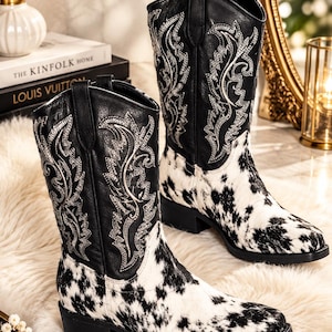 May include: A pair of black and white cow print boots featuring a black leather upper shaft with intricate white decorative stitching, a pointed toe, a low block heel, and pull tabs at the top of the shaft for a classic western-inspired silhouette.