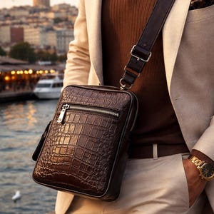 May include: A dark brown leather crossbody bag with a crocodile skin effect. The bag features a silver zip and an adjustable strap. The image shows the bag worn with a brown shirt, beige blazer, and trousers.
