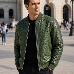 May include: A forest green bomber jacket featuring a textured ostrich leather pattern, a ribbed collar, ribbed cuffs, and a silver-toned front zipper closure. The jacket is worn over a plain black t-shirt and paired with black trousers.