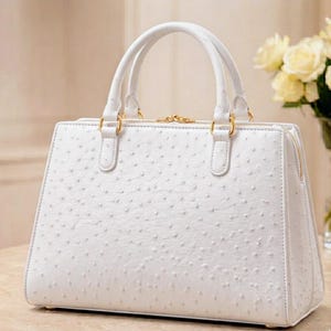 May include: A white handbag with a textured surface, featuring two top handles and gold-colored hardware. The bag has a rectangular shape with a zipper closure and a structured design. A bouquet of yellow roses is visible in the background.
