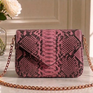 Könnte beinhalten: Rosa Crossbody-Tasche mit Schlangenmuster und goldener Kette. Die Tasche hat einen Klappenverschluss und eine rechteckige Form. Die Kette ist ca. 122 cm lang. Die Tasche liegt auf einer Marmoroberfläche.