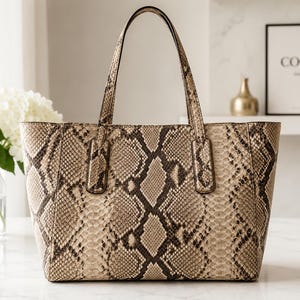 May include: A structured tote bag featuring a tan and dark brown snakeskin pattern. The handbag has two vertical shoulder straps attached to the top edge, a wide rectangular base, and a tapered silhouette with a textured, reptilian-inspired finish.