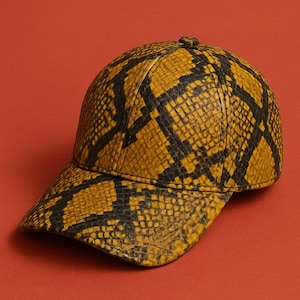 May include: A baseball cap featuring a snakeskin design in yellow and black. The cap has a curved peak and a button on the crown. The background is a solid red.