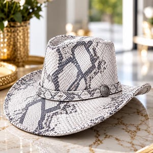 May include: A cowboy hat featuring a white and gray snakeskin pattern throughout the crown and brim. The hat includes a matching snakeskin band around the base of the crown, accented with a circular silver-toned metal concho emblem on the front.