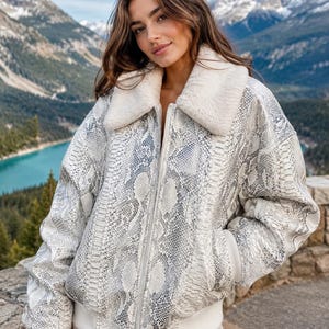 May include: A white and grey snakeskin-patterned jacket with a cream-coloured faux fur collar. The jacket has a zip closure and ribbed cuffs. The background features a mountain range and a lake.