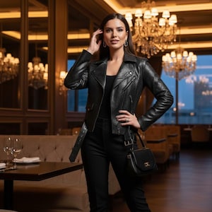 Może przedstawiać: Czarna skórzana kurtka z teksturowanym wzorem, połączona z czarnymi spodniami i małą czarną torebką. Kurtka ma pasek w talii i klasyczny styl motocyklowy. Otoczenie to restauracja z żyrandolami.