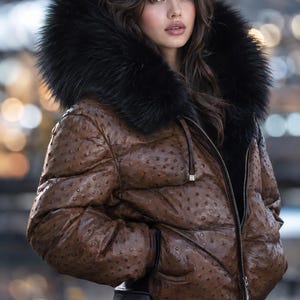 Könnte beinhalten: Eine braune Daunenjacke mit texturierter, gepunkteter Oberfläche und einer großen, schwarz gefütterten Kapuze aus Kunstfell. Die Jacke verfügt über einen Frontreißverschluss, lange Ärmel und Seitentaschen. Sie wird mit einer schwarzen Hose kombiniert.