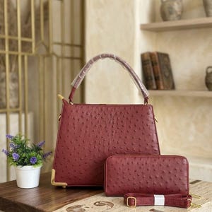 May include: A burgundy handbag and matching wallet set with a textured ostrich-like pattern. The handbag has a top handle and gold-colored corner accents. A detachable shoulder strap is included with the wallet.