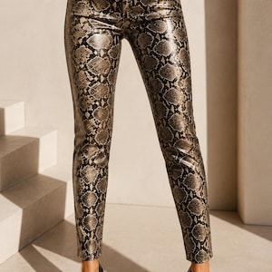 May include: High-waisted pants featuring a glossy finish and a detailed black and metallic gold snakeskin pattern. These fitted trousers include a standard waistband with a single button closure, a front zipper fly, and straight-cut legs.