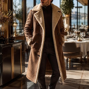 May include: A long, double-breasted brown suede coat featuring a wide, cream-coloured shearling lapel and lining. The coat has long sleeves with shearling cuffs, dark buttons, and a structured fit, worn over a dark turtleneck jumper and dark trousers.