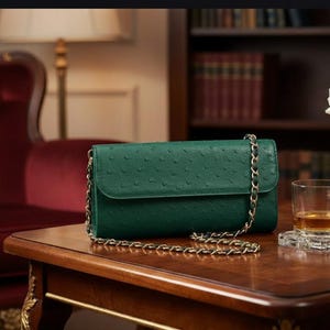 Puede incluir: Un bolso de mano verde oscuro con una correa de cadena dorada descansa sobre una mesa de madera. El bolso de mano tiene una superficie texturizada. Un vaso de líquido ámbar, una rosa blanca en un jarrón dorado y un par de gafas también están sobre la mesa.