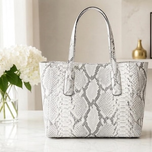 May include: A structured tote bag featuring a white and gray snakeskin pattern. The handbag has two vertical handles attached to the top edge, a rectangular base, and a wide opening. The exterior surface displays a detailed reptilian scale texture.