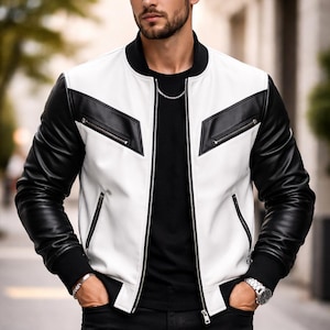 Puede incluir: Chaqueta bomber de cuero blanco y negro con cuello y puños negros. La chaqueta tiene cierre de cremallera y dos bolsillos delanteros. Las mangas son negras y el cuerpo es blanco con detalles negros. La persona lleva una camiseta negra y pantalones negros.