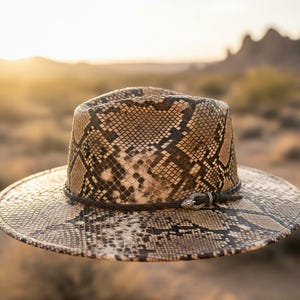 Könnte beinhalten: Ein Fedora-Hut mit Schlangenmuster und breiter Krempe. Der Hut hat ein schwarzes Band um den Hutkrempe und einen silberfarbenen Zierverschluss. Der Hut ist in den Farben Beige, Braun und Schwarz gehalten.